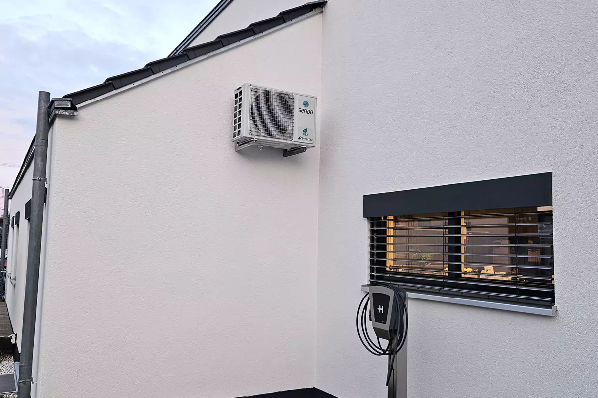 Wandmontierte Klimaanlage neben einem Fenster mit Jalousien und einer Ladestation für Elektrofahrzeuge.